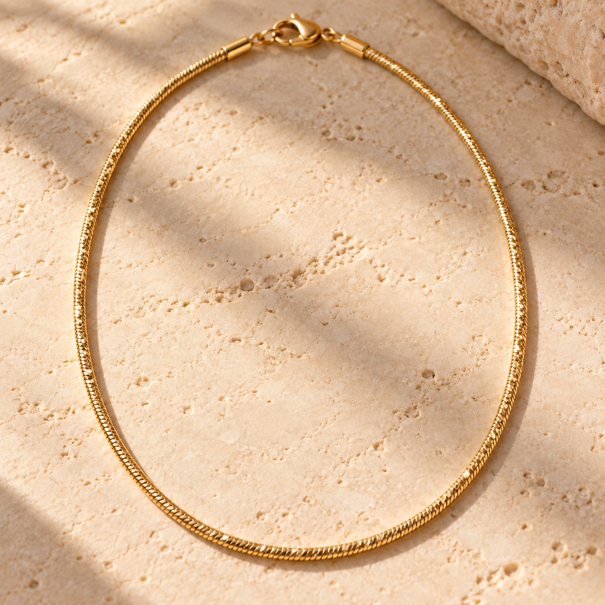 Bar None diamond-cut gold snake chain necklace on travertine, facets catching the light